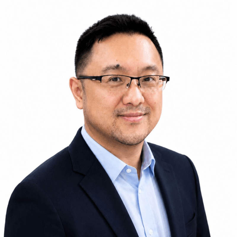 Professional headshot of a man in a navy suit and light blue shirt, wearing glasses, looking at the camera on a white background.